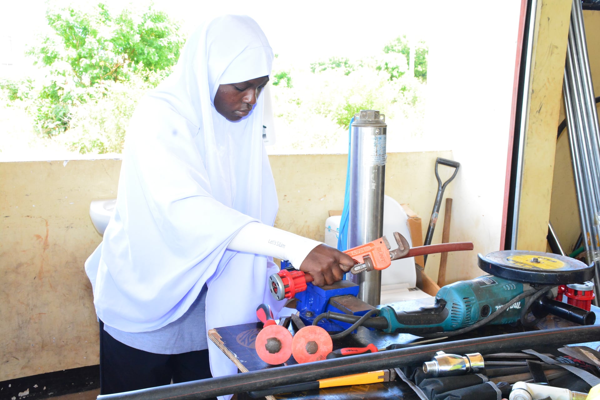 FDC student in a white hijab gripping a heavy pipe wrench at a plumbing workbench